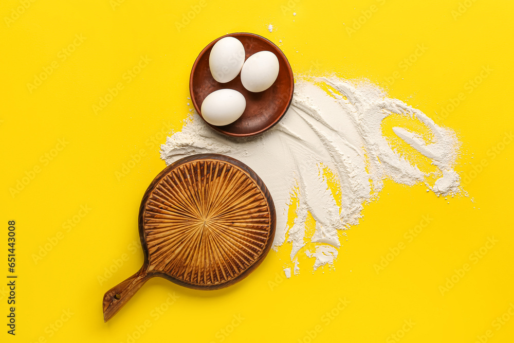 Composition with wooden cutting board, chicken eggs and flour on yellow background