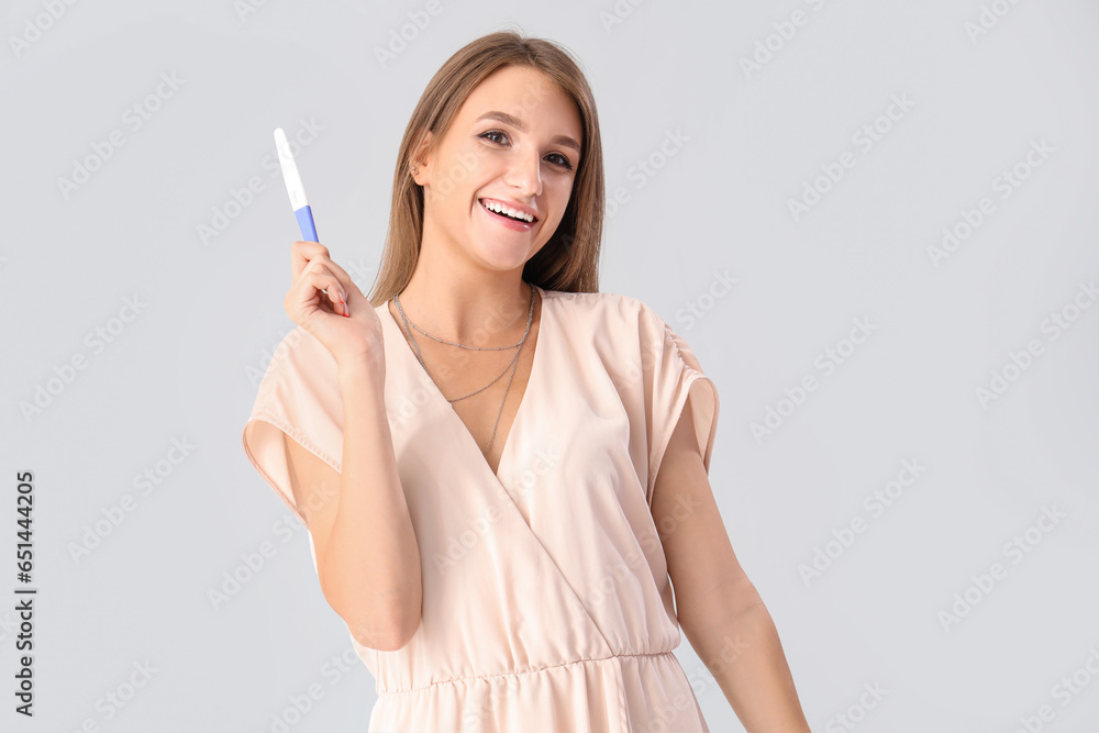 Happy beautiful young woman with pregnancy test on grey background