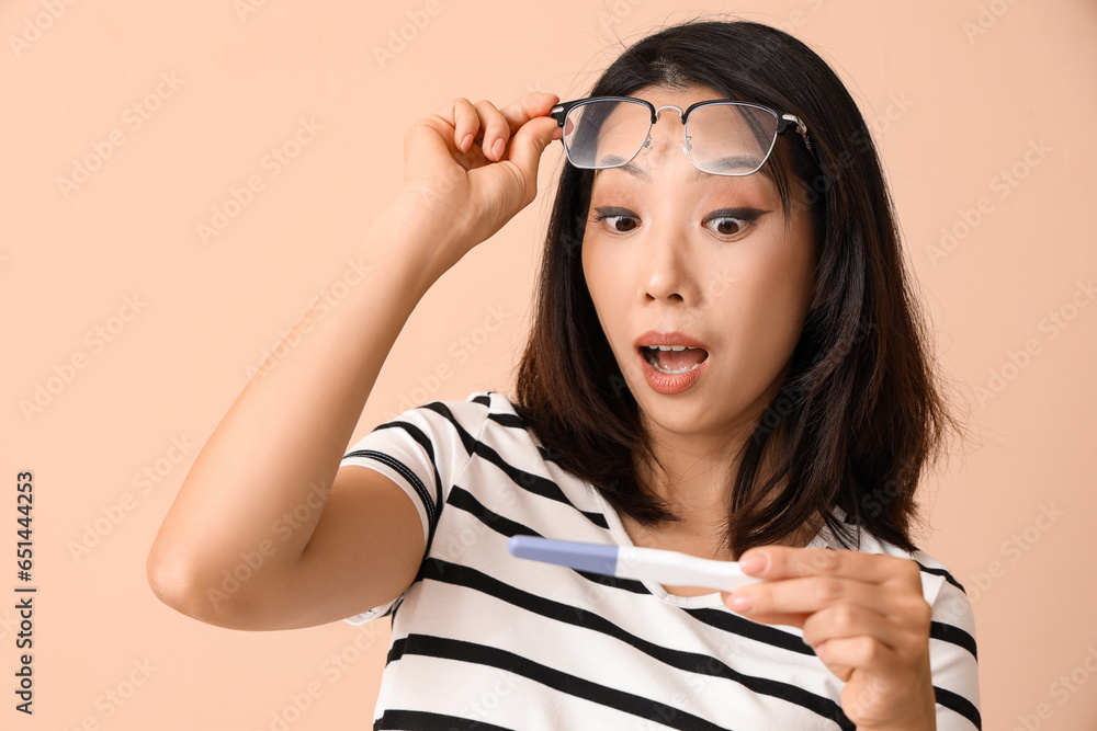 Shocked beautiful young Asian woman with pregnancy test on beige background