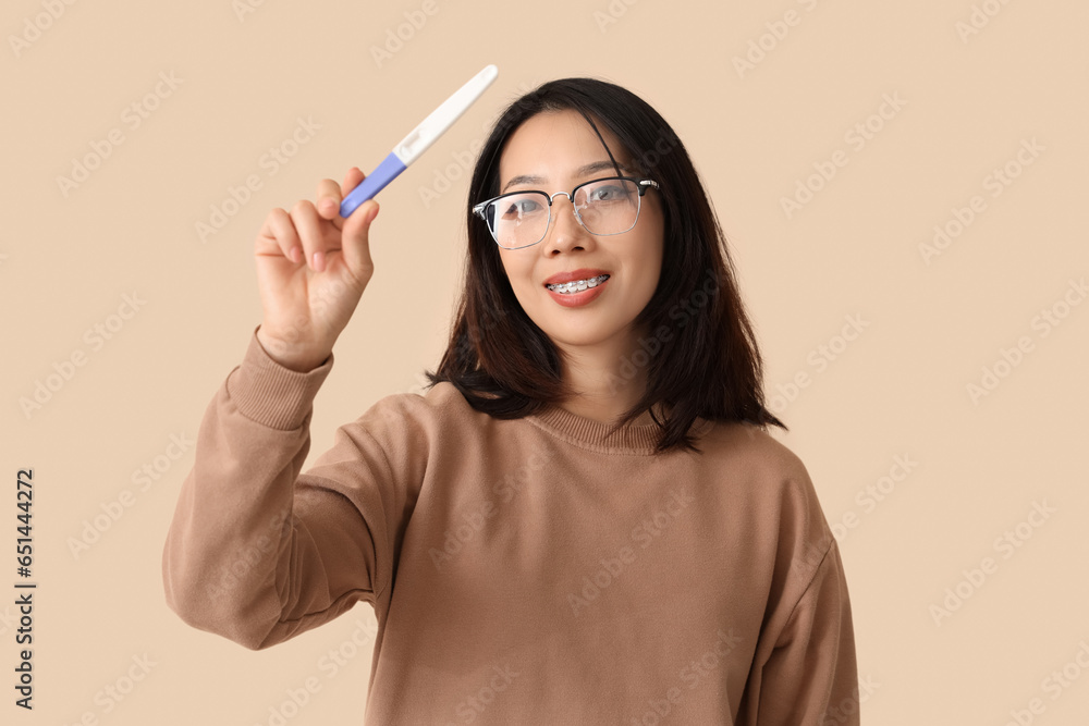 Happy beautiful young Asian woman with pregnancy test on beige background