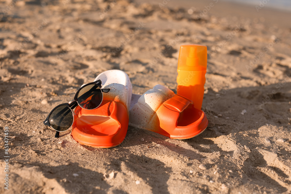 Stylish female flip flops, bottle of sunscreen and sunglasses on sand