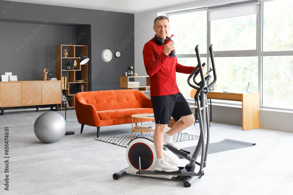 Sporty mature man with bottle of water training on elliptical trainer at home