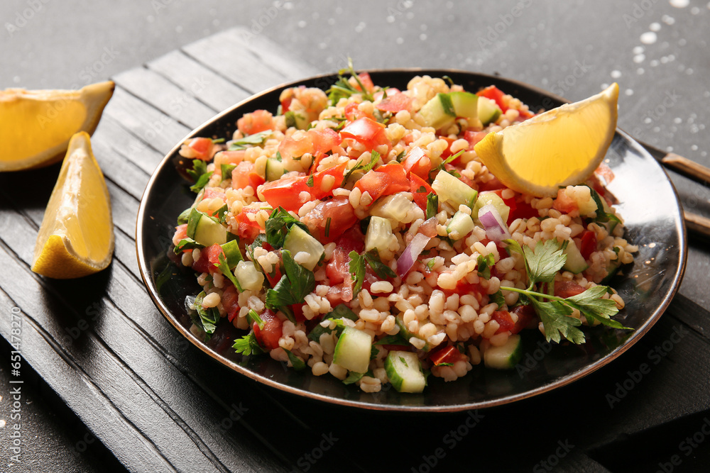 Plate with delicious tabbouleh salad and lemon pieces on dark background, closeup