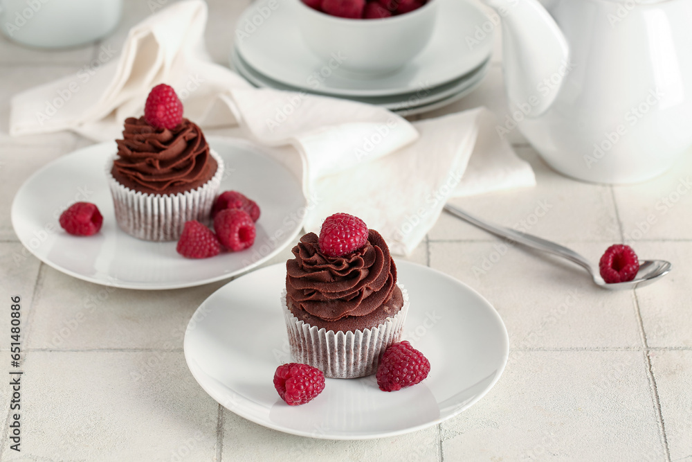 Plates of tasty chocolate cupcakes with raspberries on white tile background