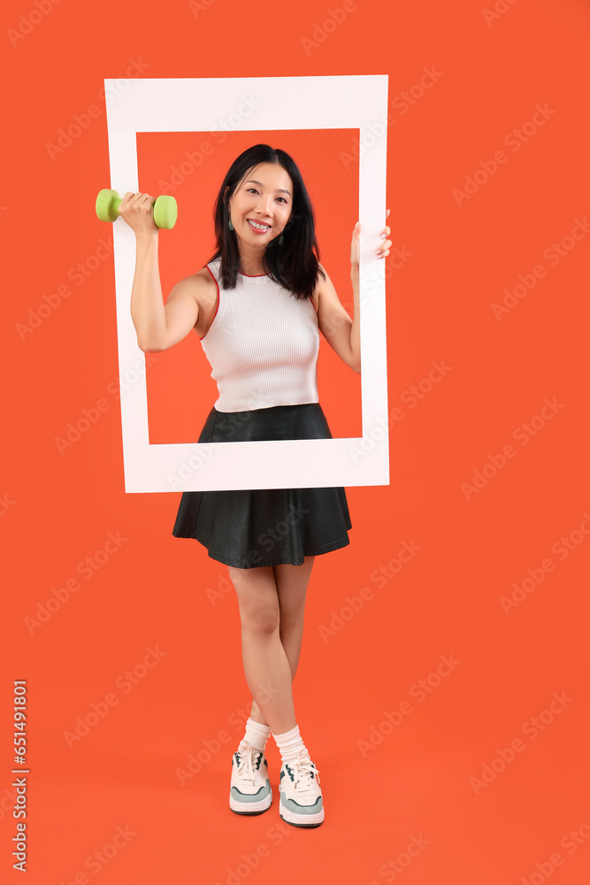 Young beautiful Asian woman with frame and dumbbell on orange background