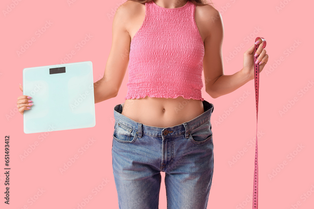 Beautiful young woman in loose jeans with measuring tape and scales on pink background. Weight loss concept