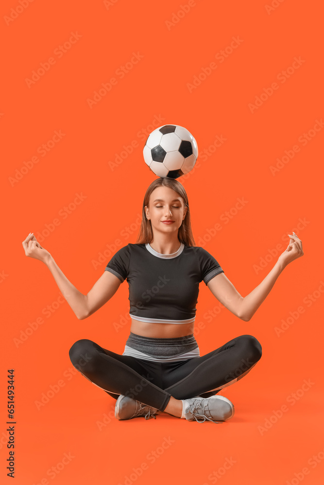 Beautiful young sporty woman with soccer ball meditating on orange background