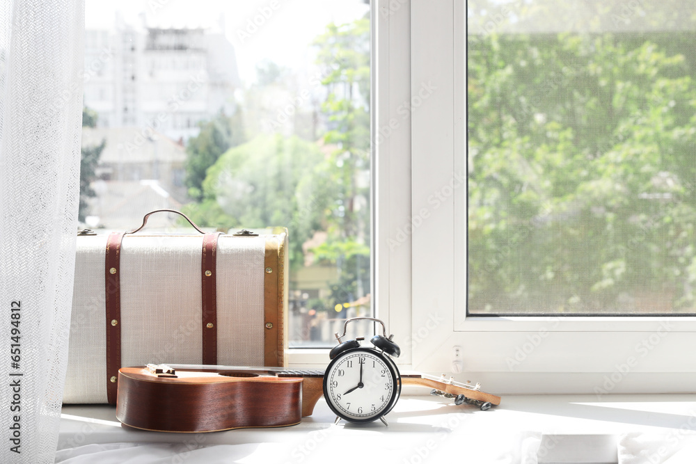 Alarm clock, ukulele and suitcase on windowsill in bedroom