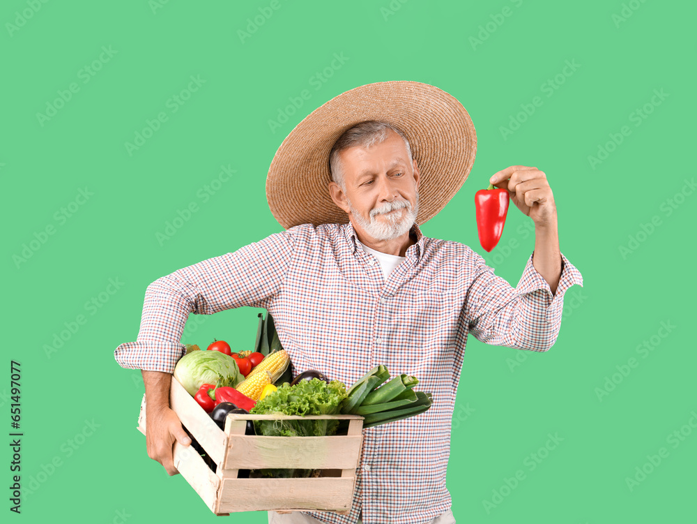 Mature male farmer with pepper and wooden box full of different ripe vegetables on green background