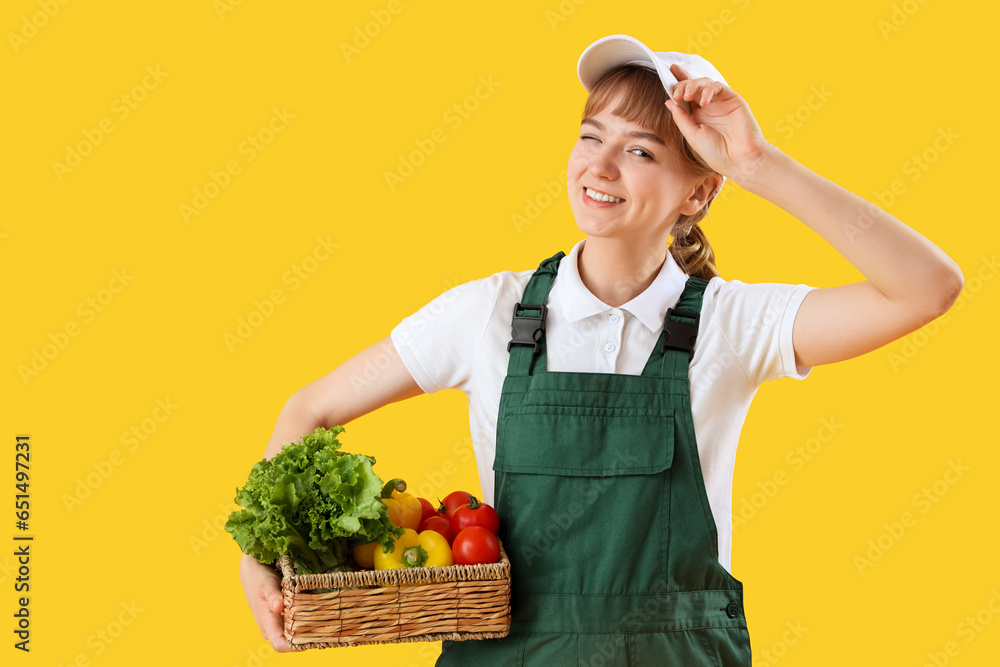 Young female farmer with wicker basket full of different ripe vegetables on yellow background