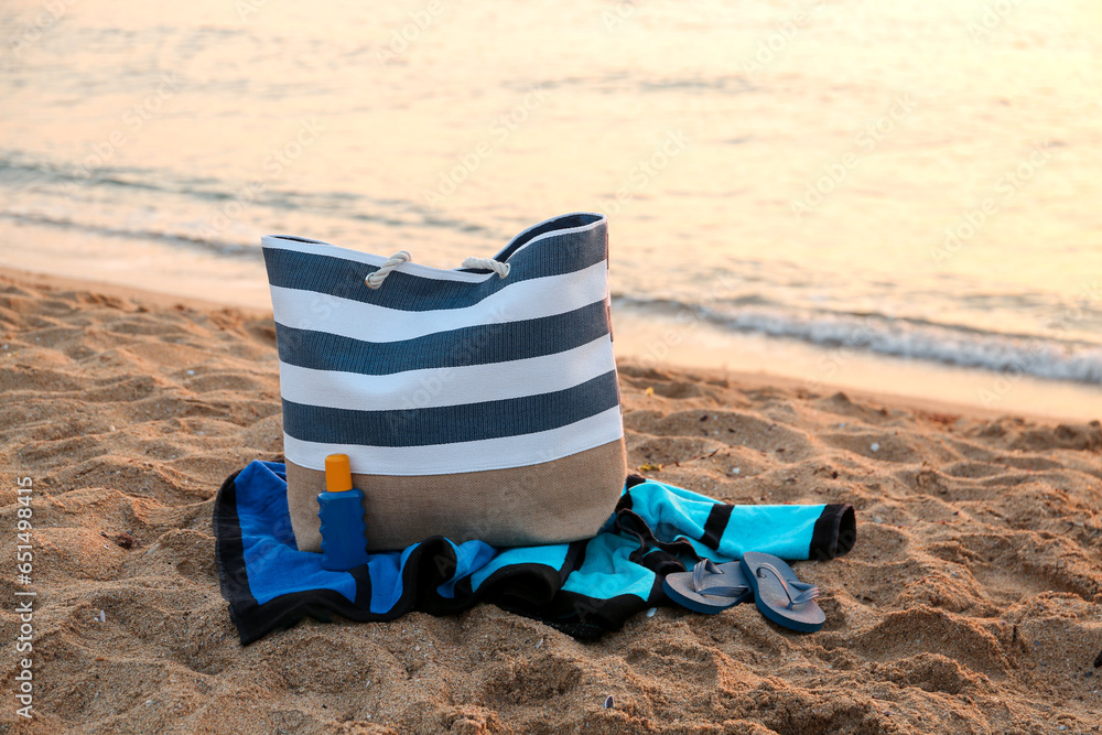 Stylish beach bag, flip-flops, sunscreen cream and towel on sand near sea