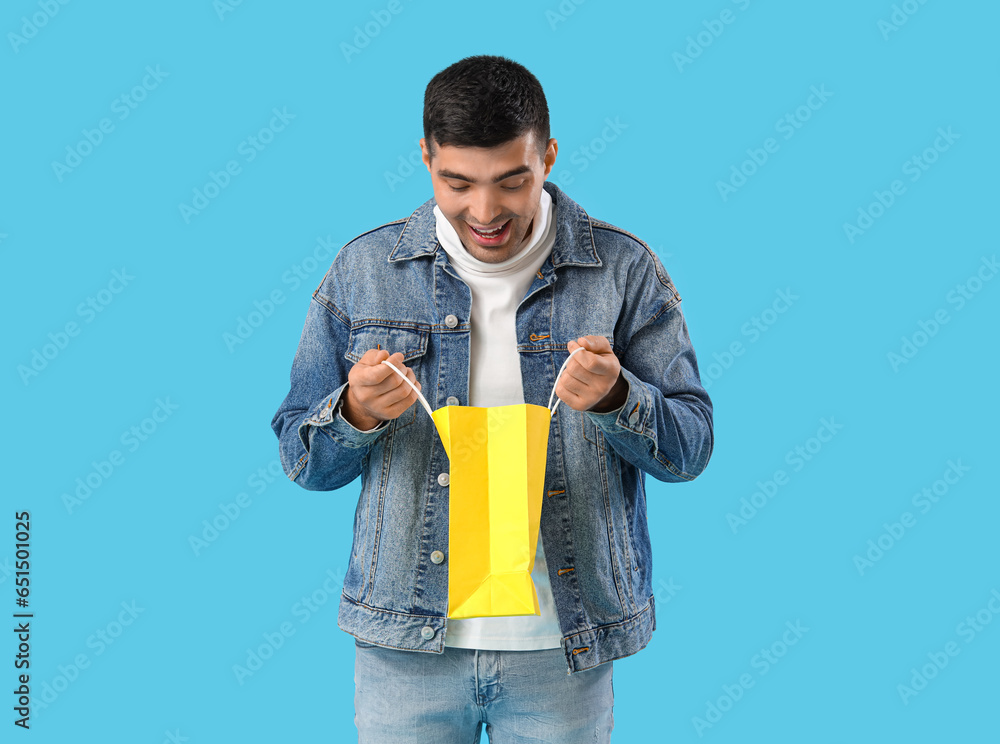 Surprised handsome young man opening shopping bag on blue background