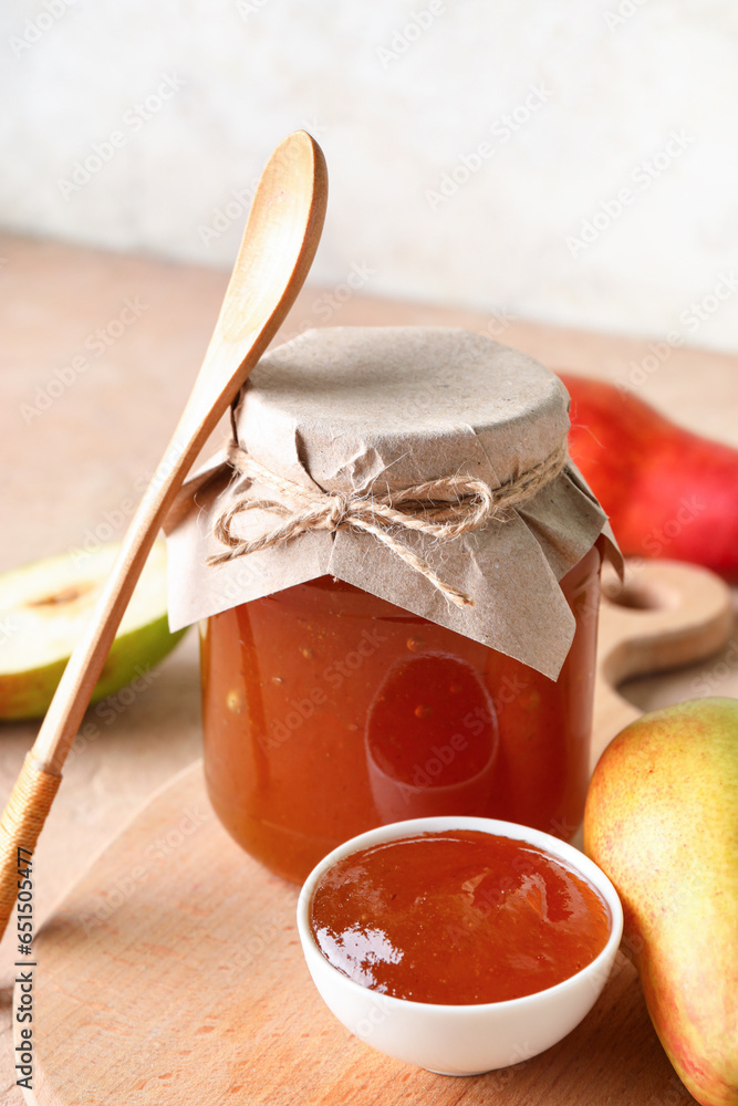 Glass jar and bowl of tasty pear jam with wooden board on table