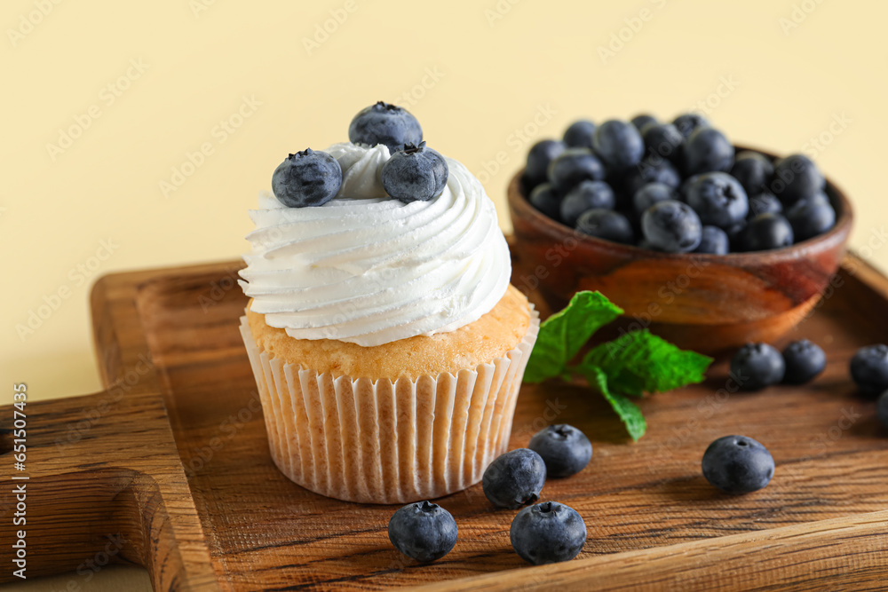 Wooden board of tasty cupcake with blueberries on yellow background