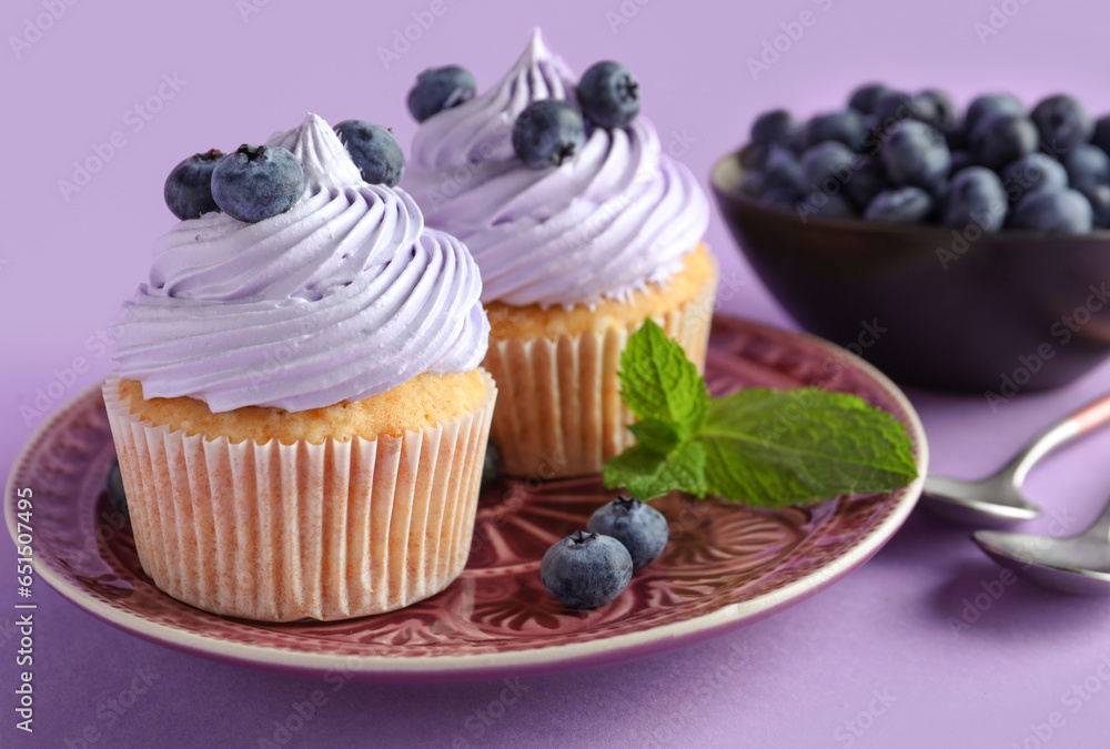 Plate of tasty cupcakes with blueberries on lilac background