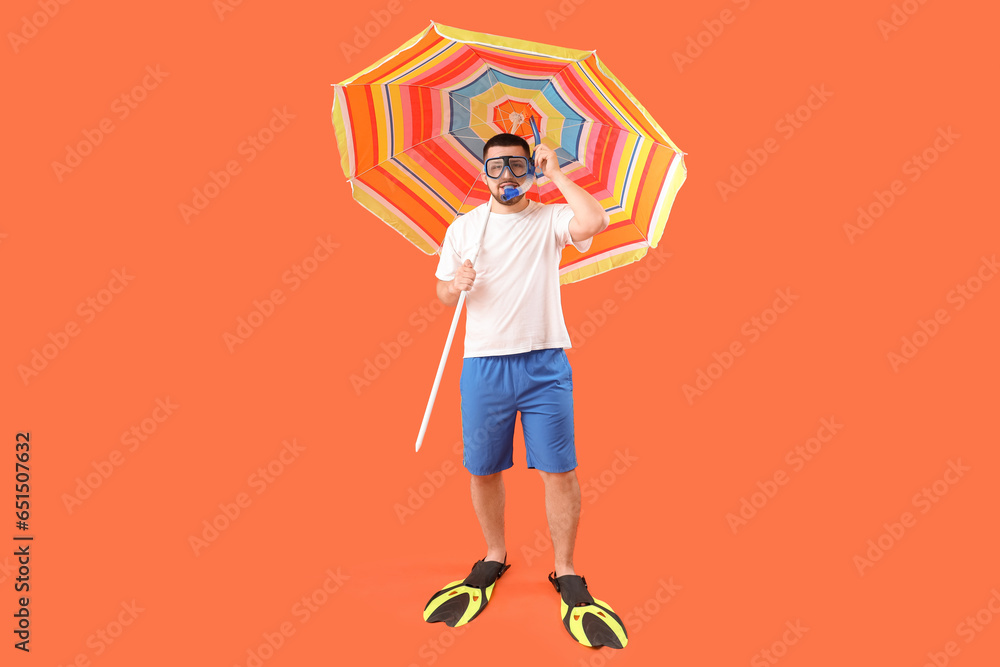 Handsome young man with flippers, snorkeling mask and umbrella on red background