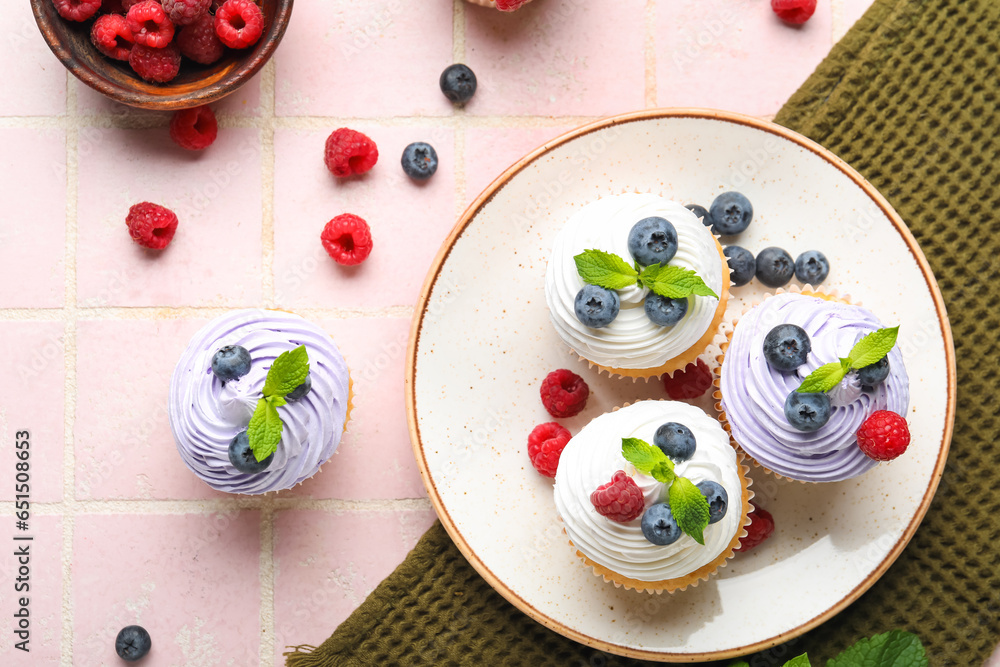 Plate of delicious cupcakes with blueberries, raspberries and mint on pink tile background