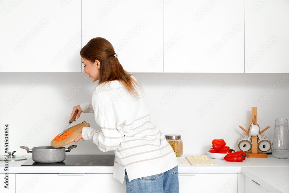 Young woman cooking chicken soup with cut carrot on stove in kitchen