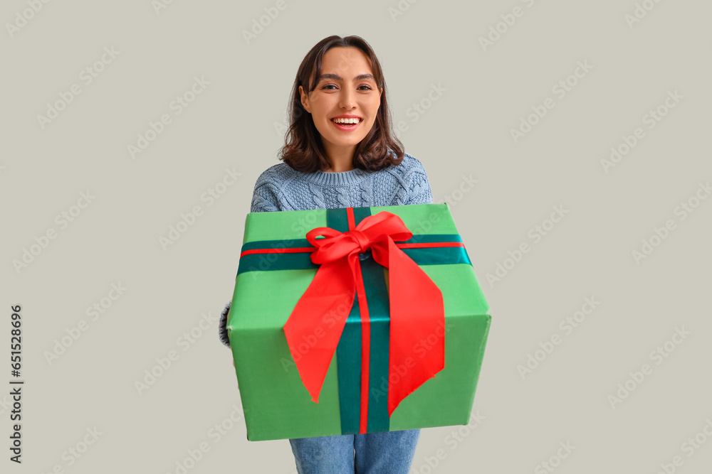 Young woman in warm sweater with Christmas gift on light background