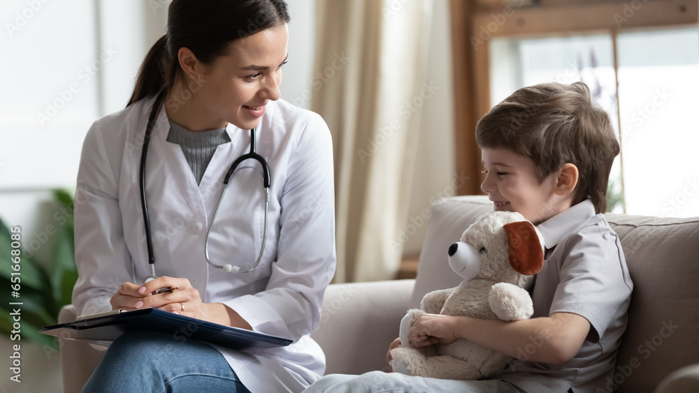 Friendly female doctor consulting little patient at meeting, writing notes in card, laughing positive preschool child hugging teddy toy, talking to smiling pediatrician, children healthcare concept