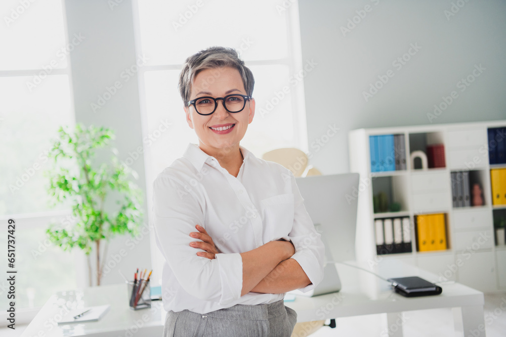 Photo of confident positive lady employer dressed white shirt eyewear arms folded smiling indoors workstation workshop