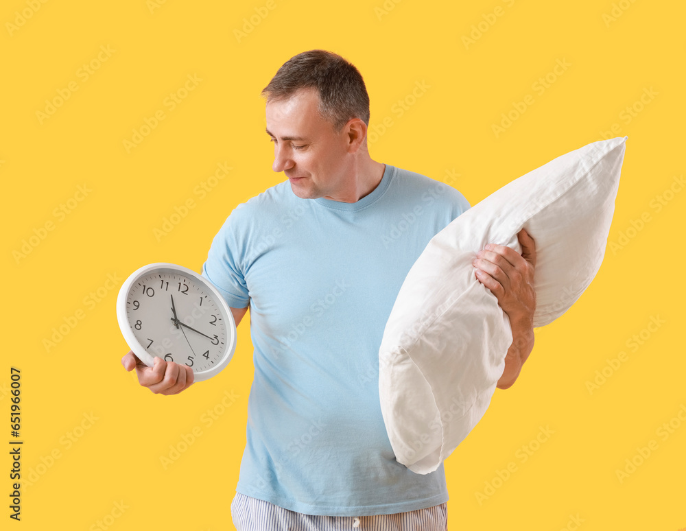 Mature man with soft pillow and clock on yellow background