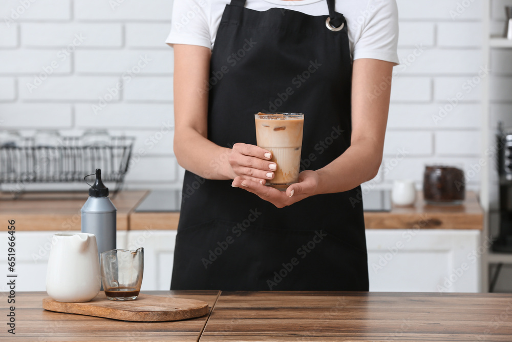 Female barista holding glass of tasty coffee on wooden table in kitchen