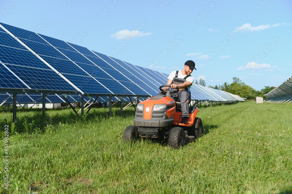 A man working at solar power station. A worker on a garden tractor mows grass on a solar panel farm.