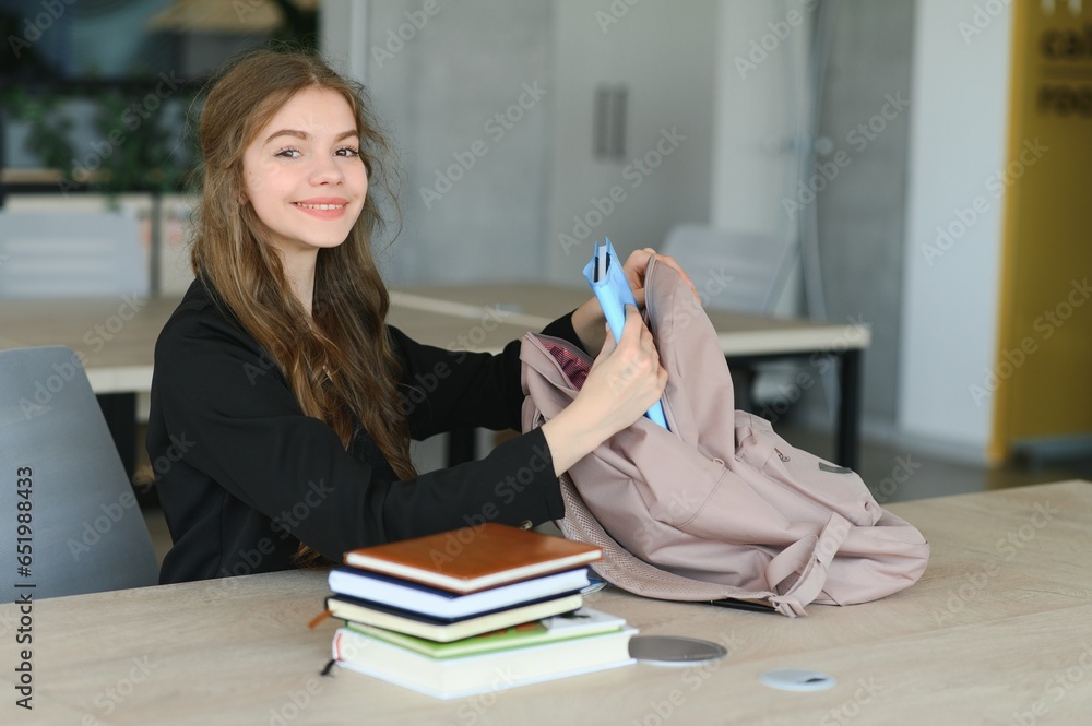 A cute 15-year-old schoolgirl sits at a school desk with notebooks and books and studies