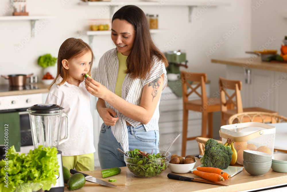 Little girl with her mother eating cucumber in kitchen