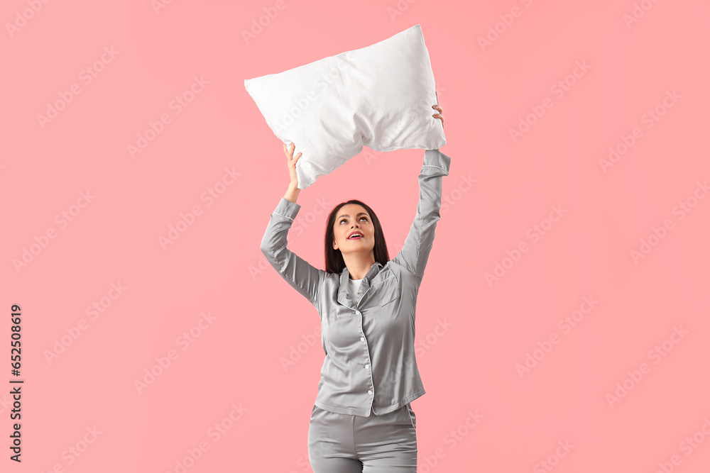 Pretty young woman in pajamas and with pillow on pink background