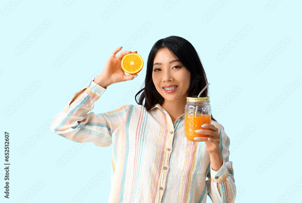 Beautiful Asian woman with glass jar of juice and orange on blue background