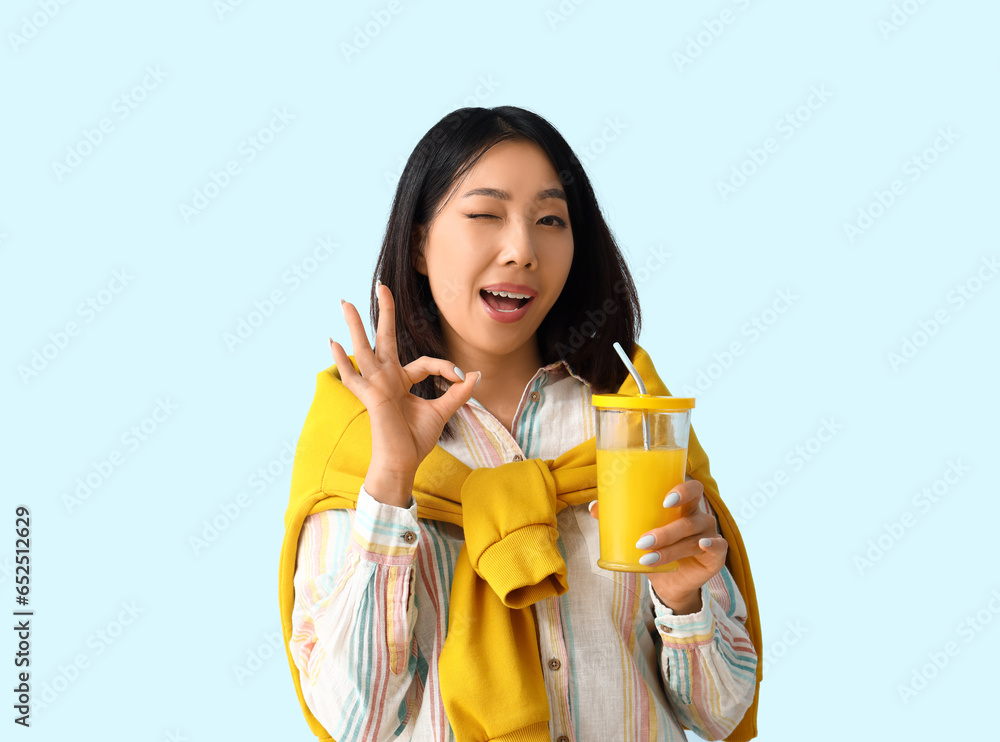 Beautiful Asian woman with glass of juice showing OK on blue background