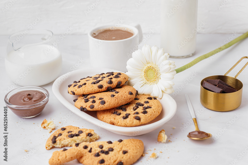 Plate of tasty cookies with chocolate chips, milk, sauce, cup of coffee and chamomile flower on white background