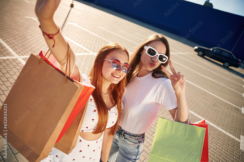 Taking a selfie. Two beautiful women in casual clothes are holding shopping bags, outdoors