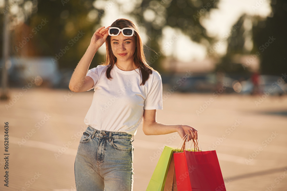 Wearing the sunglasses, standing. Beautiful woman in casual clothes is holding shopping bags, outdoors