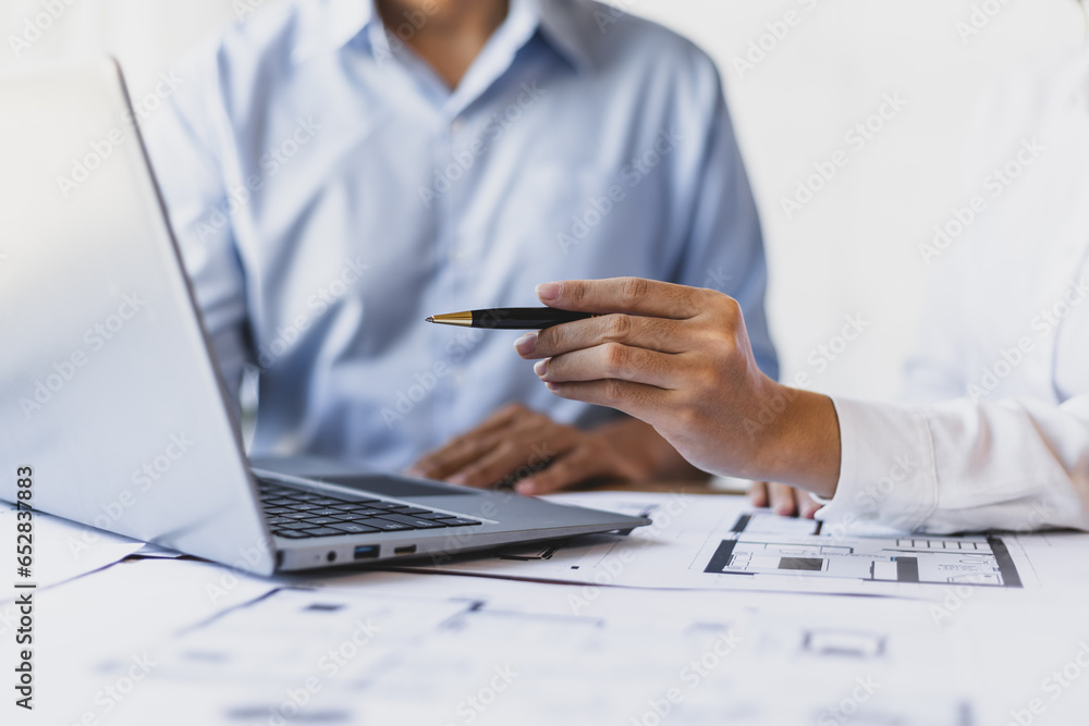 Brainstorming group of architects and engineers discuss building blueprint design on laptop and construction project planning on wooden table in office.