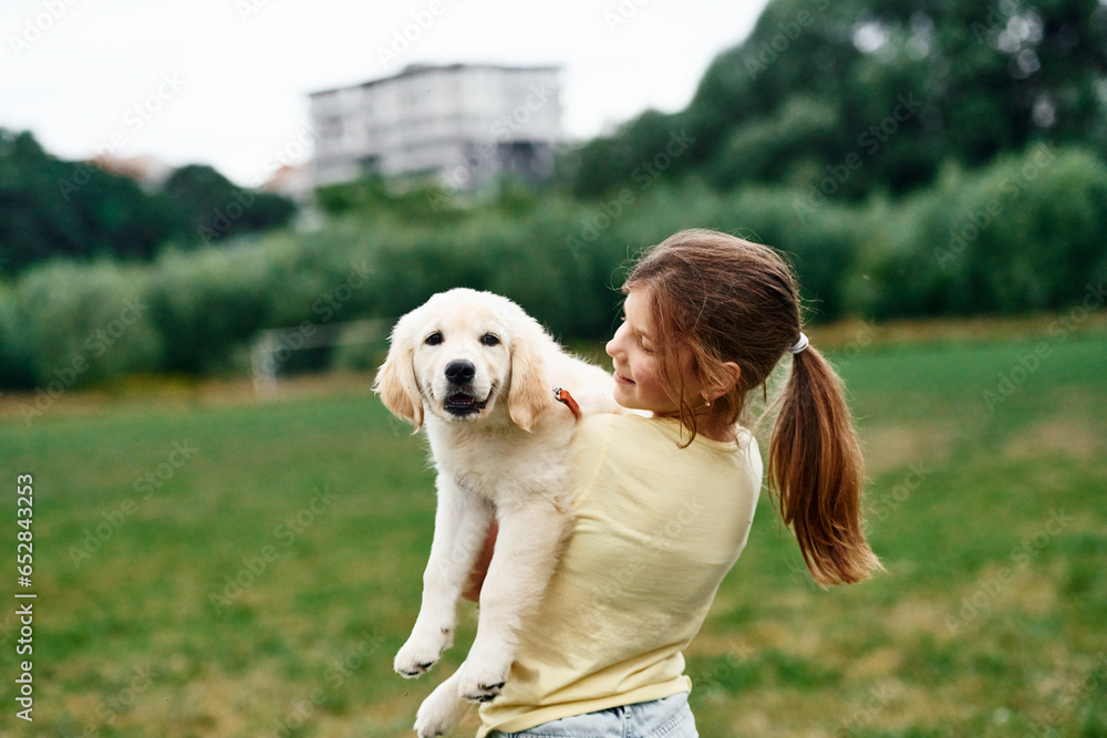 Holding animal in hands. Cute little girl is with golden retriever dog on the field