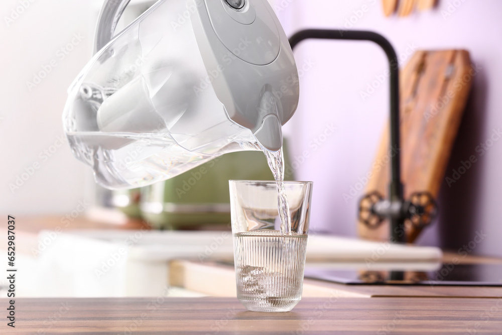 Pouring of water from modern filter jug into glass on kitchen counter, closeup