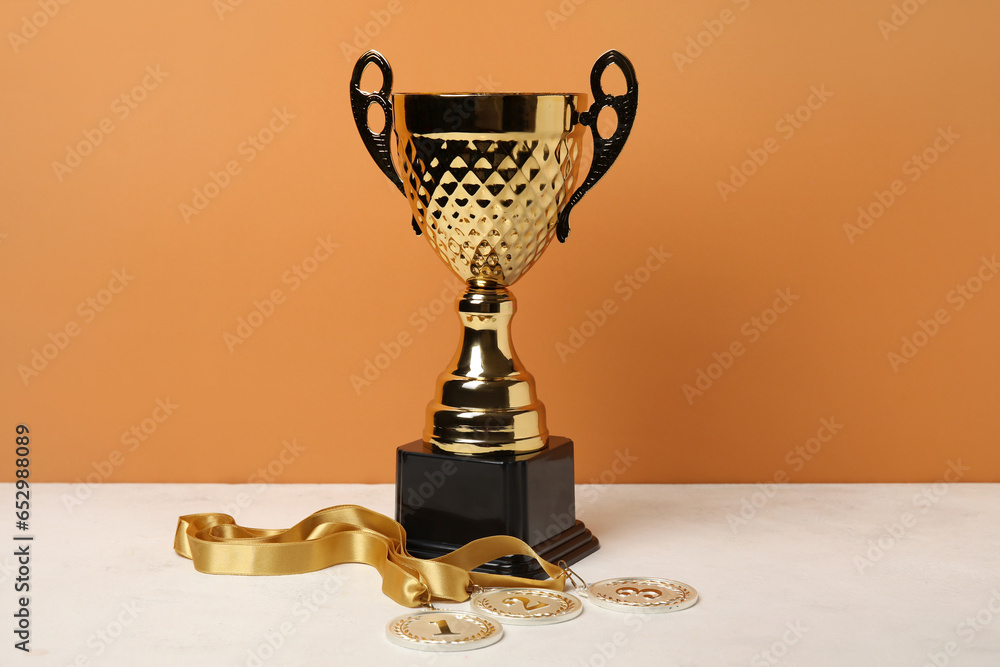 Gold cup with prize medals on table near orange wall