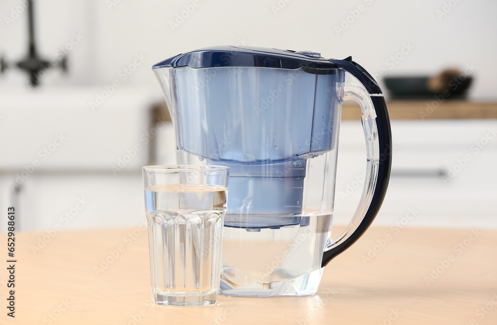 Glass of pure water and filter jug on wooden table in kitchen