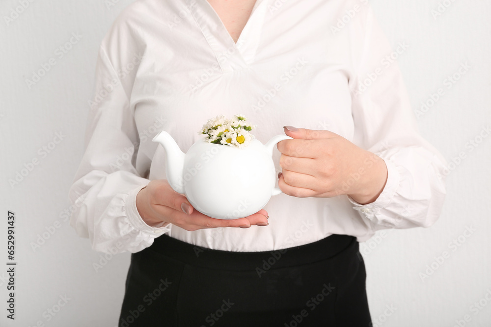 Beautiful young woman with teapot of chamomile tea and flowers near white wall