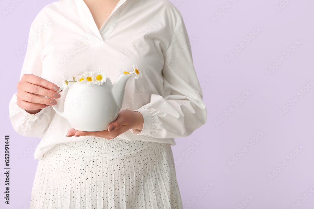 Beautiful young woman with teapot of chamomile tea and flowers near lilac wall