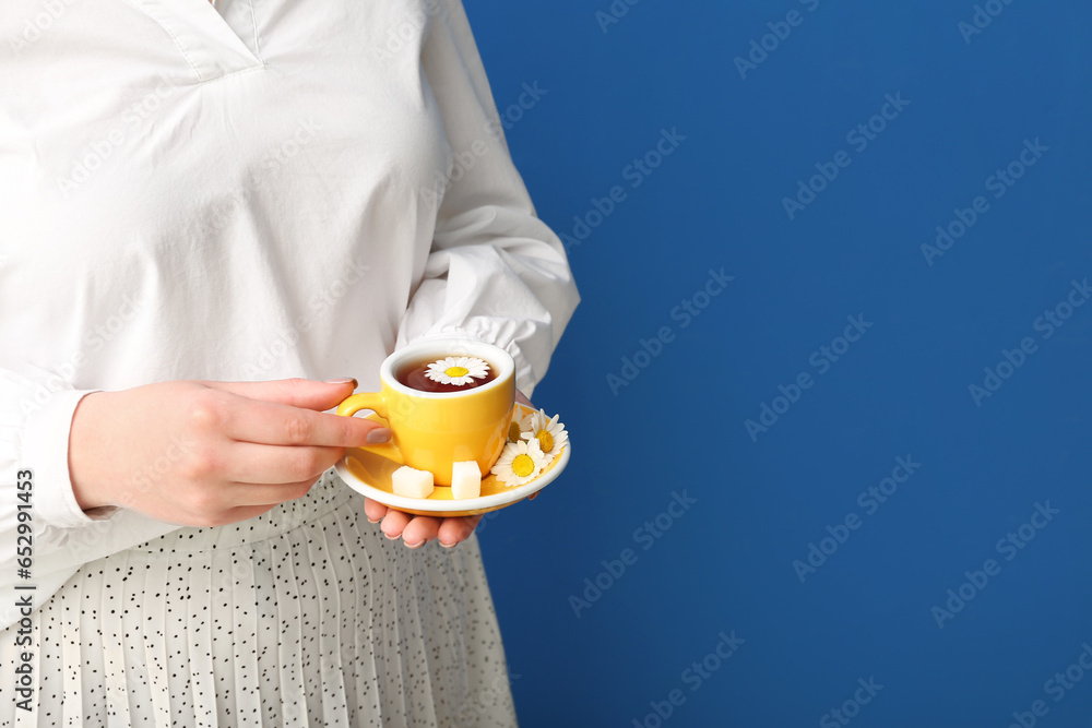 Beautiful young woman with cup of chamomile tea and flowers near blue wall