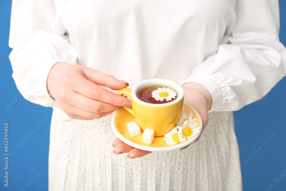 Beautiful young woman with cup of chamomile tea and flowers near blue wall