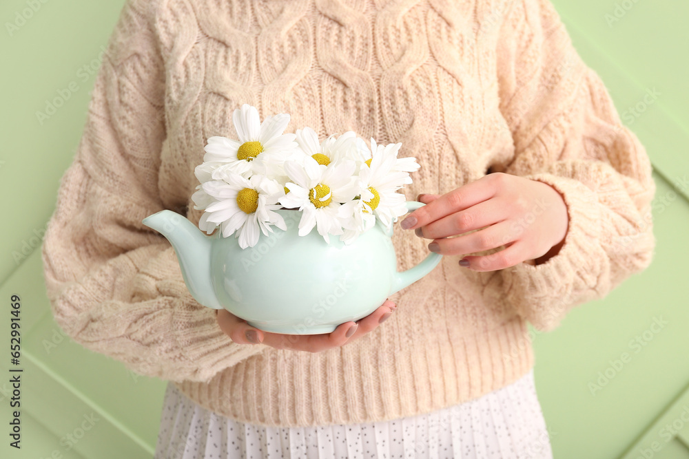 Beautiful young woman with teapot of chamomile tea and flowers near green wall