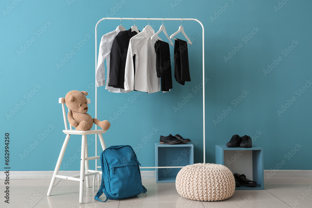 Stylish school uniform hanging on rack, backpack, shoes and pouf near blue wall in room