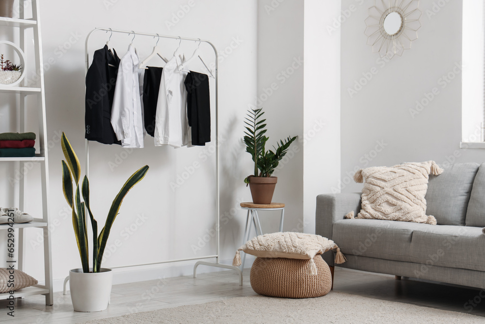 Interior of light living room with stylish school uniform hanging on rack, shelving unit, sofa and houseplants