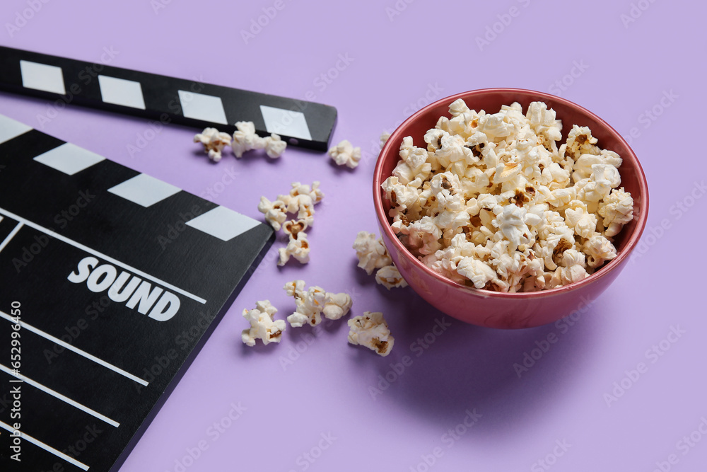 Bowl with tasty popcorn and clapperboard on purple background