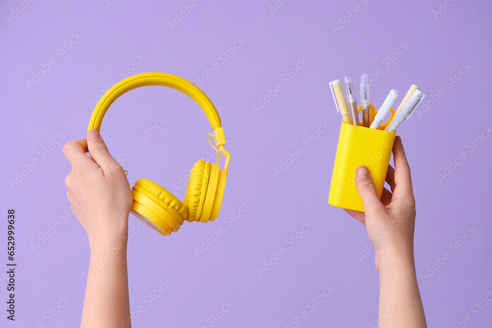 Female hands holding headphones and cup with pens on lilac background